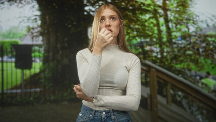 Obraz premium Woman with hand to mouth and crossed arm by wooden railing on park walkway wearing cream sweater and jeans; thoughtful solitude.