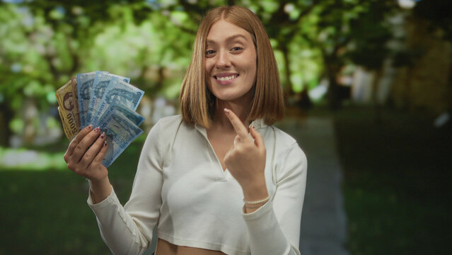 Young blonde woman in a park holding hungarian forint banknotes outdoors, expressing happiness and financial success, vibrant greenery in the background highlighting economic prosperity.