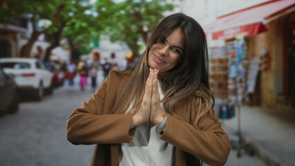 Fototapeta premium Woman smiling in brown coat with hands in prayer gesture on a lively city street with trees and shops in the background suggesting a vibrant urban atmosphere.