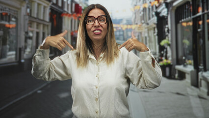 Obraz premium Woman pointing both fingers at her chest wearing large glasses and white shirt on a street; confidence empowerment.