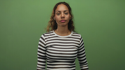 Fototapeta premium Woman points index finger upward in studio wearing striped shirt against green backdrop; confidence focus.