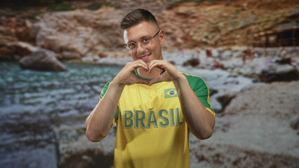 Naklejka premium Man wearing yellow brasil jersey and glasses smiling hands under chin beside rocky river shore in forest setting; pride.
