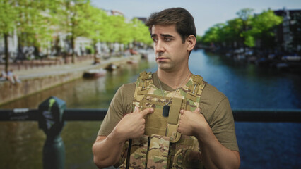 Man soldier points to dogtag while wearing camouflage vest on street by canal bridge; duty...