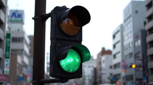 Close‑up of a traffic light with green signal illuminated, mounted on a pole in an urban Japanese setting with multi‑story buildings in the background under an overcast sky
