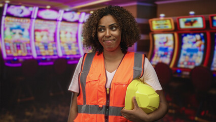 Obraz premium Engineer woman holding yellow hardhat and wearing high visibility orange vest beside slot machines in casino building; confidence professionalism.