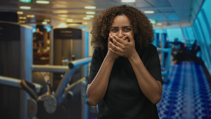 Fototapeta premium Woman with hands covering mouth and gym machines visible in building, smiling with wide eyes and ring on finger; surprise.