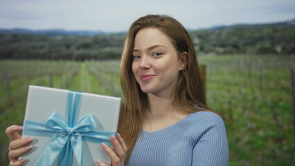 Obraz premium Woman smiles and holds wrapped gift box with blue ribbon in green vineyard outdoors; celebration anticipation joy.