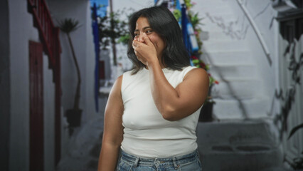 Fototapeta premium Young hispanic woman in white sleeveless top and jeans covers mouth with hand while smiling on a narrow street by white steps; surprise delight.