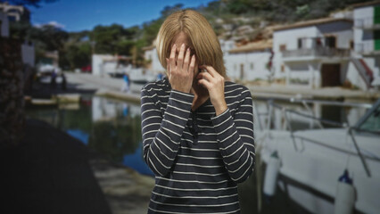 Fototapeta premium Blonde woman in striped top holding smartphone to ear while covering her face with one hand on street by marina; anxiety.