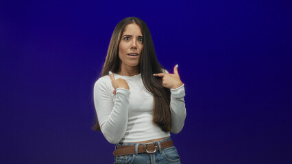 Fototapeta premium Young woman wearing white long sleeve shirt and jeans displays palms out stop gesture and points thumb to chest in studio; hesitation doubt.