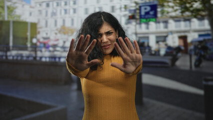 Fototapeta premium Young arab brunette woman clutching chest for heartattack on busy city street wearing mustard sweater and visible hand piercings; emotional distress compassion.