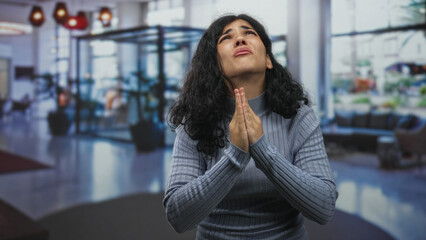 Woman with hands clasped in prayer looking upward in a hotel building lobby, visible pleading...