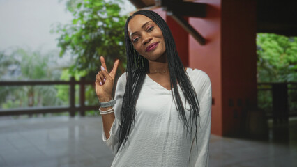 Fototapeta premium Woman showing peace sign with hand and holding small lipgloss near face in building wearing white blouse and braided hair; playful confidence.