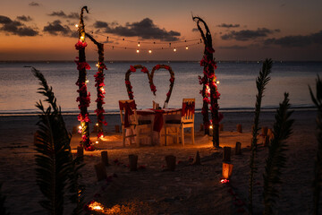 A romantic dinner table for two set on a beach at sunset with flower arches and decorative lights. Location: Zanzibar, Tanzania.