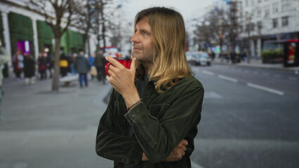 Fototapeta premium Blond man sipping from red ceramic coffee cup on busy street lined with buildings; contemplation serenity.