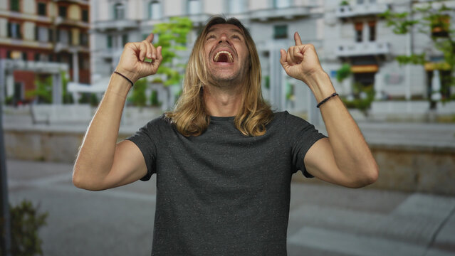 Man with long blond hair pointing both index fingers upward on urban street while smiling broadly in daylight; optimism.