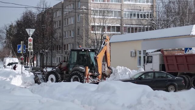 Heavy machinery removing accumulated snow from urban street. Backhoe loader with front attachment plowing snow, creating large piles. Dump truck parked nearby.