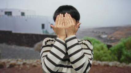 Woman outdoors playfully covering her face and sticking out tongue, wearing striped sweater with...