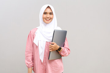 Portrait of a smiling Asian woman wearing a hijab, looking forward while holding a laptop, isolated on a gray background.