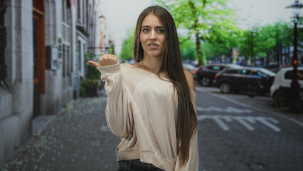 Obraz premium Teenage girl with long hair and braces points thumb backward while wearing off shoulder sweater on tree lined city street with parked cars and visible crosswalk; uncertainty.