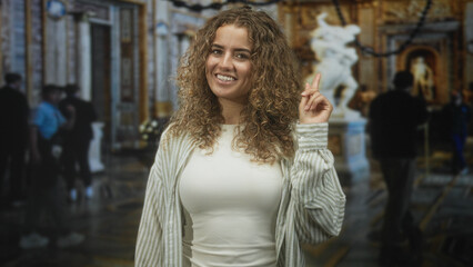 Fototapeta premium Woman points finger up and smiles, finger raised inside ornate museum building with marble statue and visitors; curiosity wonder.