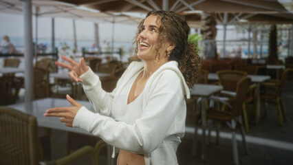 Fototapeta premium Young woman with curly hair smiles and gestures with hands, showing navel piercing in an outdoor seaside cafe by the boardwalk; joyful carefree.