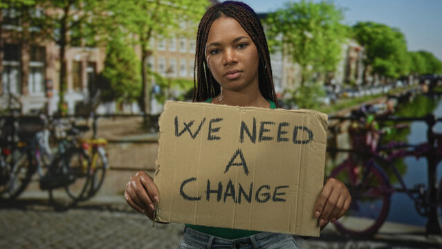 Woman holding cardboard sign reading we need a change on amsterdam canal street with parked bicycles and canal water visible; determination.