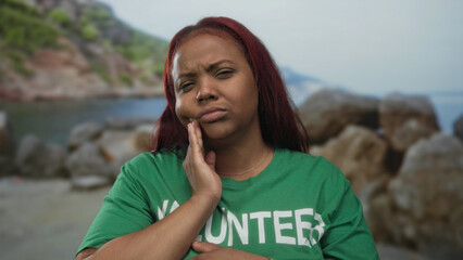 Volunteer woman holding cheek with palm gesture stands at a sunny beach shore showing discomfort  toothache. © Krakenimages.com