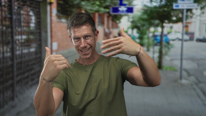Naklejka premium Man wearing glasses gesturing with hands and smiling on a busy city street near brick building; friendly invitation.