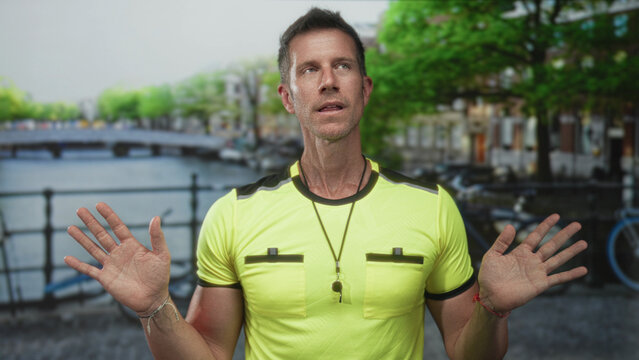 Man referee in neon yellow shirt with whistle, hands raised palms out on street in amsterdam; confidence.