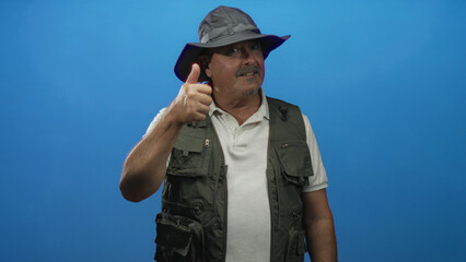 Man wearing fishing vest and wide brim hat shows thumbs up with right hand and looks sideways in studio; adventure confidence.