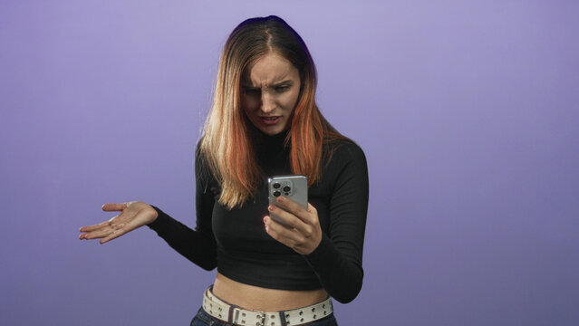 Young redhead woman holding smartphone, hands gripping device and frowning at screen while wearing black crop top and studded belt in studio; frustration tech problem.