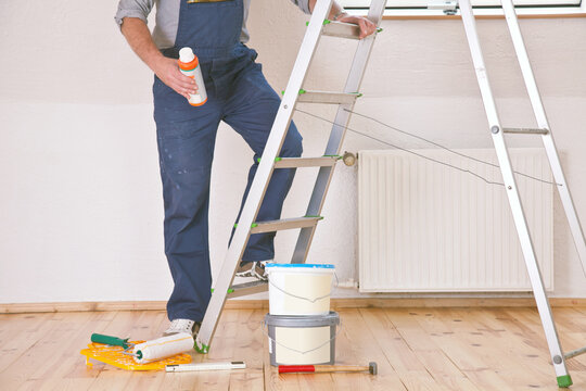 Handwerker im Arbeitskleidung bei Malerarbeiten vor einer Leiter mit verschiedenen Farbeimern und Werkzeugen.