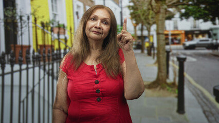 Fototapeta premium Senior hispanic woman in red dress pointing index finger upward on a street with iron railings and trees; reflective advice.