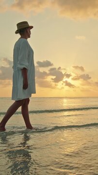 Young woman walking barefoot in shallow sea at sunset and sunrise on Lipa Noi beach, Koh Samui, peaceful tropical travel moment with golden sky 