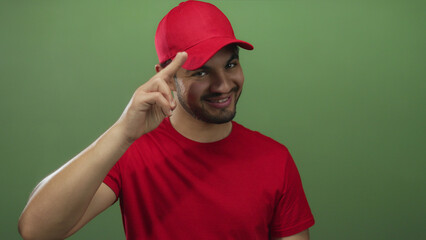 Young delivery courier man in red shirt salutes his red cap with fingers in studio setting; respect...