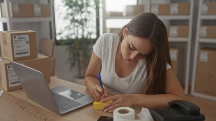 Woman writing on sticky note with pen at desk in office with stacked parcels and laptop visible;...