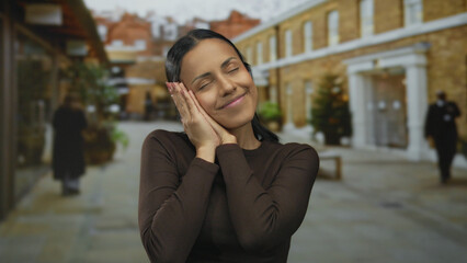 Fototapeta premium Young hispanic woman smiling peacefully with closed eyes on a quiet outdoor street with background buildings and trees, embodying serenity and relaxation.