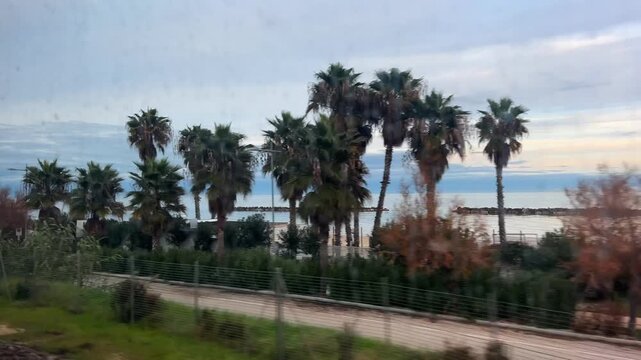 Seaside coastline at sunset seen through the window of a moving train. Reflections on the glass. Waves, breakwater rocks, and tourist facilities such as bars and resorts pass by in the foreground