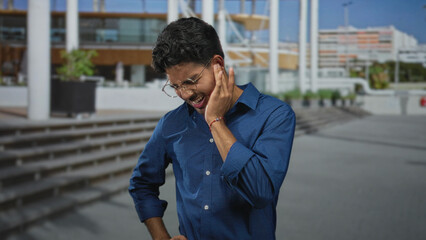 Fototapeta premium Man holding cheek with hand, wearing glasses and blue shirt, wincing near public building steps and plaza entrance; discomfort reflection.