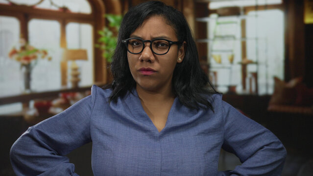 Woman in blue blouse with glasses and furrowed brow stands arms akimbo in building lit by daylight; annoyance.