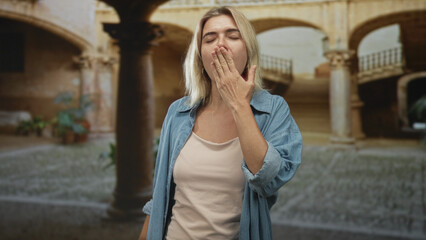 Fototapeta premium Woman yawning with hand covering mouth in stone building courtyard, denim shirt and beige tanktop visible; fatigue.