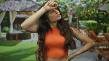 Obraz premium Woman in orange top with long brown hair shields forehead with hand in forest; summer heat exhaustion.