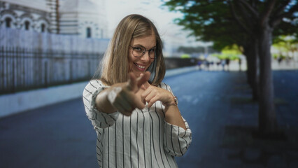 Fototapeta premium Woman pointing finger to camera in urban street with blurred building facade and tree lined walkway; confidence.