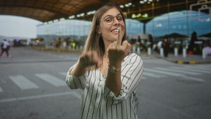 Fototapeta premium Blonde woman flashing middle finger and miming a crank motion outside an airport terminal arrivals area; defiance insult.