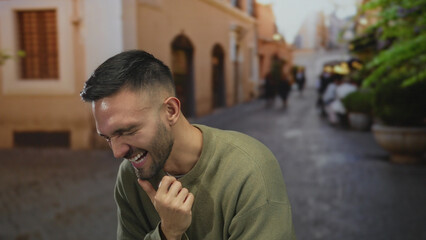 Obraz premium Young hispanic man joyfully laughing outdoors on a charming urban city street.