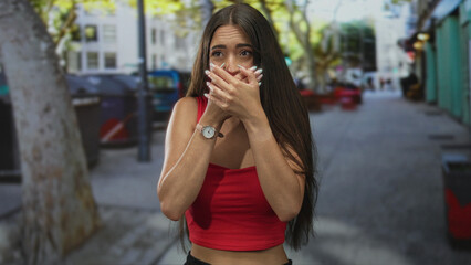 Fototapeta premium Young hispanic woman with long hair and hands covering mouth on a busy city street, wearing red crop top and wristwatch; surprise silence.