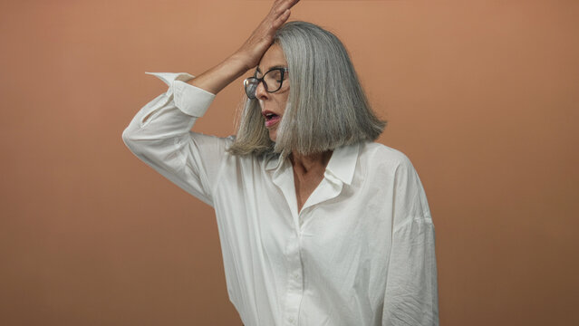 Woman with gray hair holds hand to forehead in studio with peach backdrop and white shirt; surprise realization.