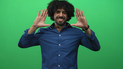Fototapeta premium Man with hands up, palms facing camera and smiling, curly hair visible against a green studio backdrop; playful joy.