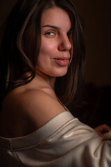 Fototapeta premium Young woman with long dark hair wearing an oversized white sweater poses seated against a textured wall in a softly lit indoor setting
